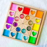 A wooden Montessori-style sorting puzzle board with colorful translucent gem-like pieces arranged in a grid on a white quilted blanket.