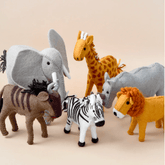 A set of handcrafted felt safari animal toys including an elephant, giraffe, rhinoceros, lion, wildebeest, and zebra, displayed against a neutral background.