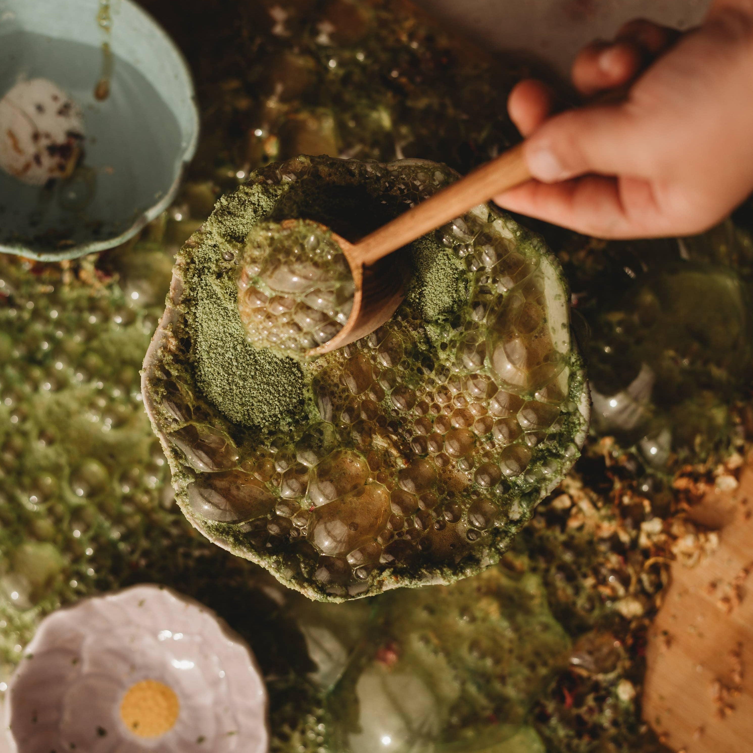 Person holding a wooden spoon over a bowl filled with green liquid in a rustic setting.