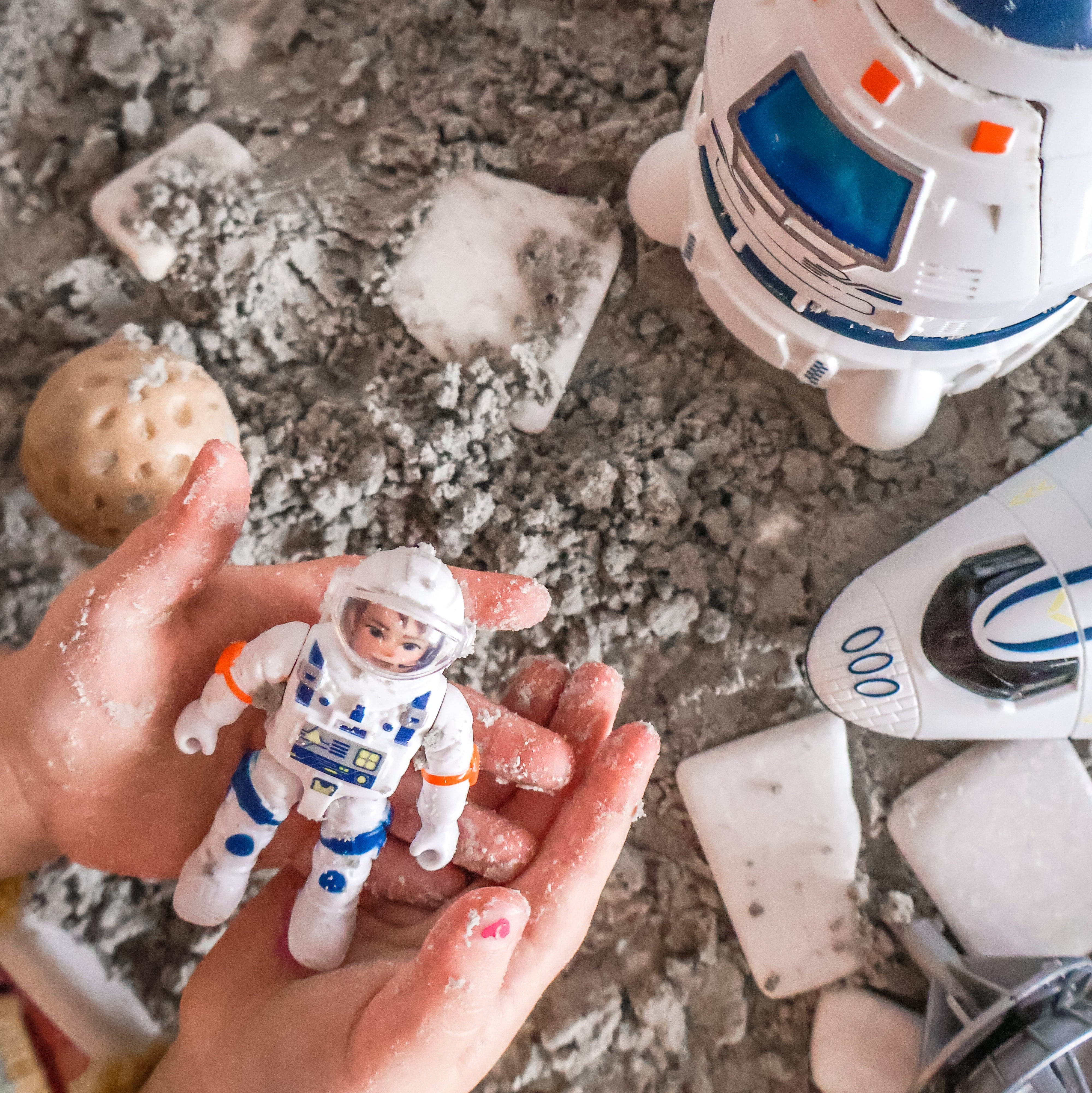 Toy astronaut figure held by a hand on a sandy surface with nature sensory stone and vehicle in the background.