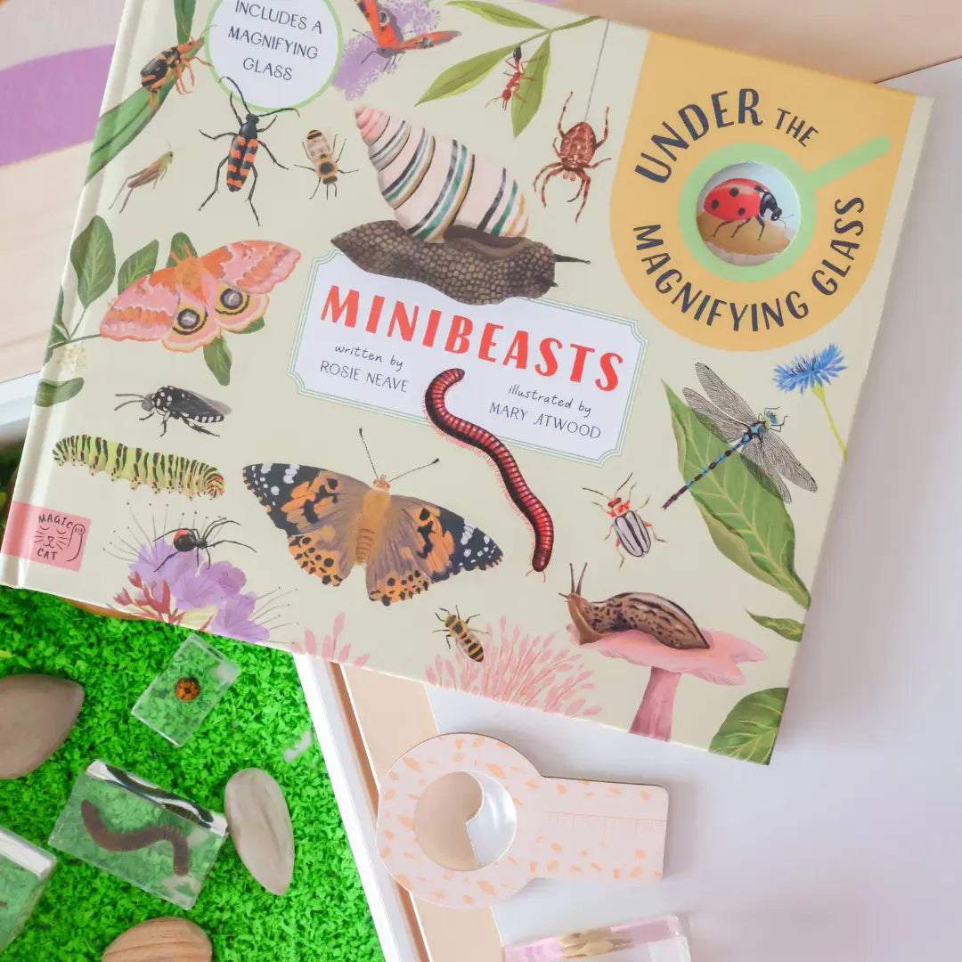 The book 'Minibeasts: Under the Magnifying Glass' by Rosie Naylor and Mark Jernigan, featuring colorful insect and creature illustrations on a cream cover, styled on a white surface next to a green mossy tray with mini magnifying glass accessories.
