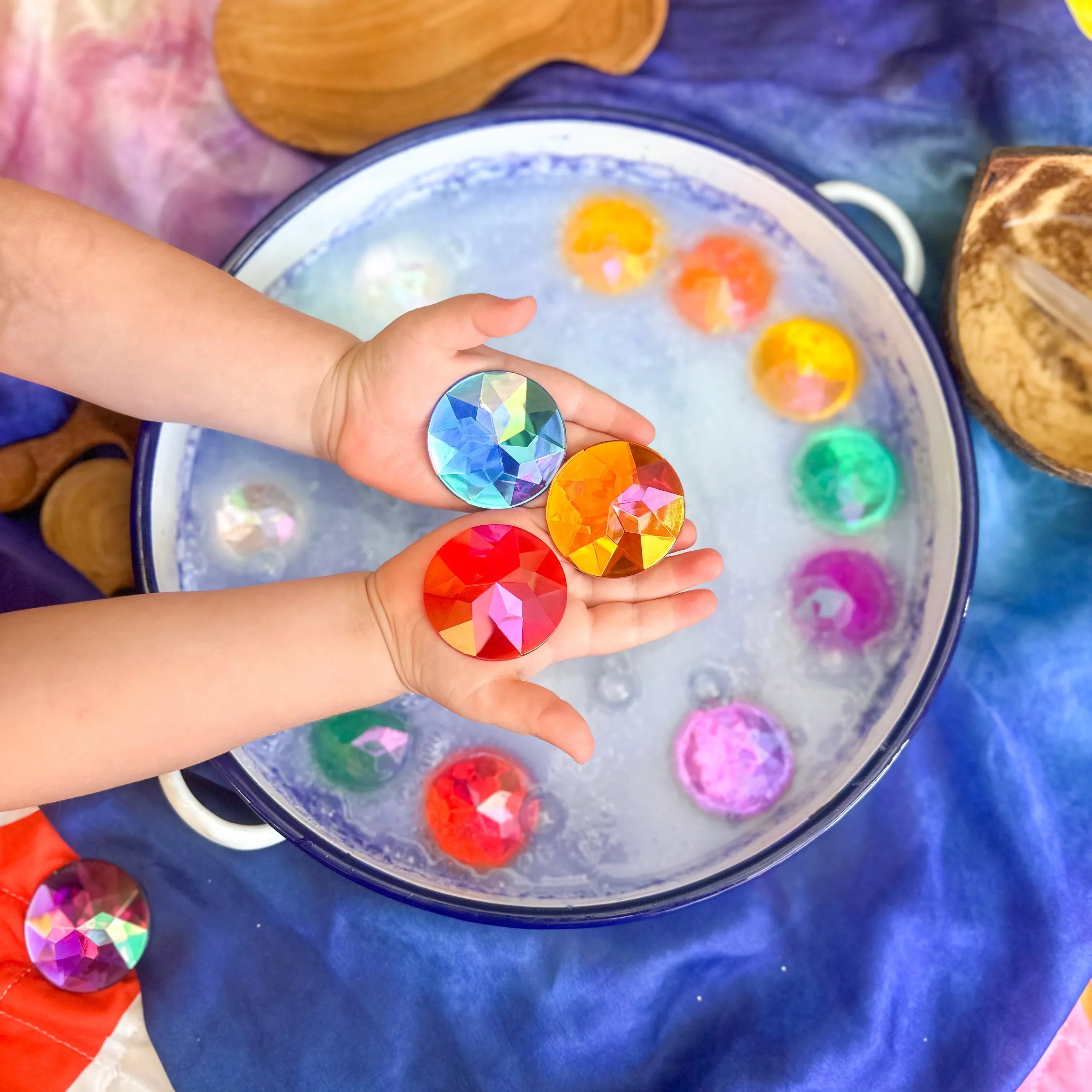 Child's hands holding colorful gemstones over a water play setup with bubbles and wooden toys.