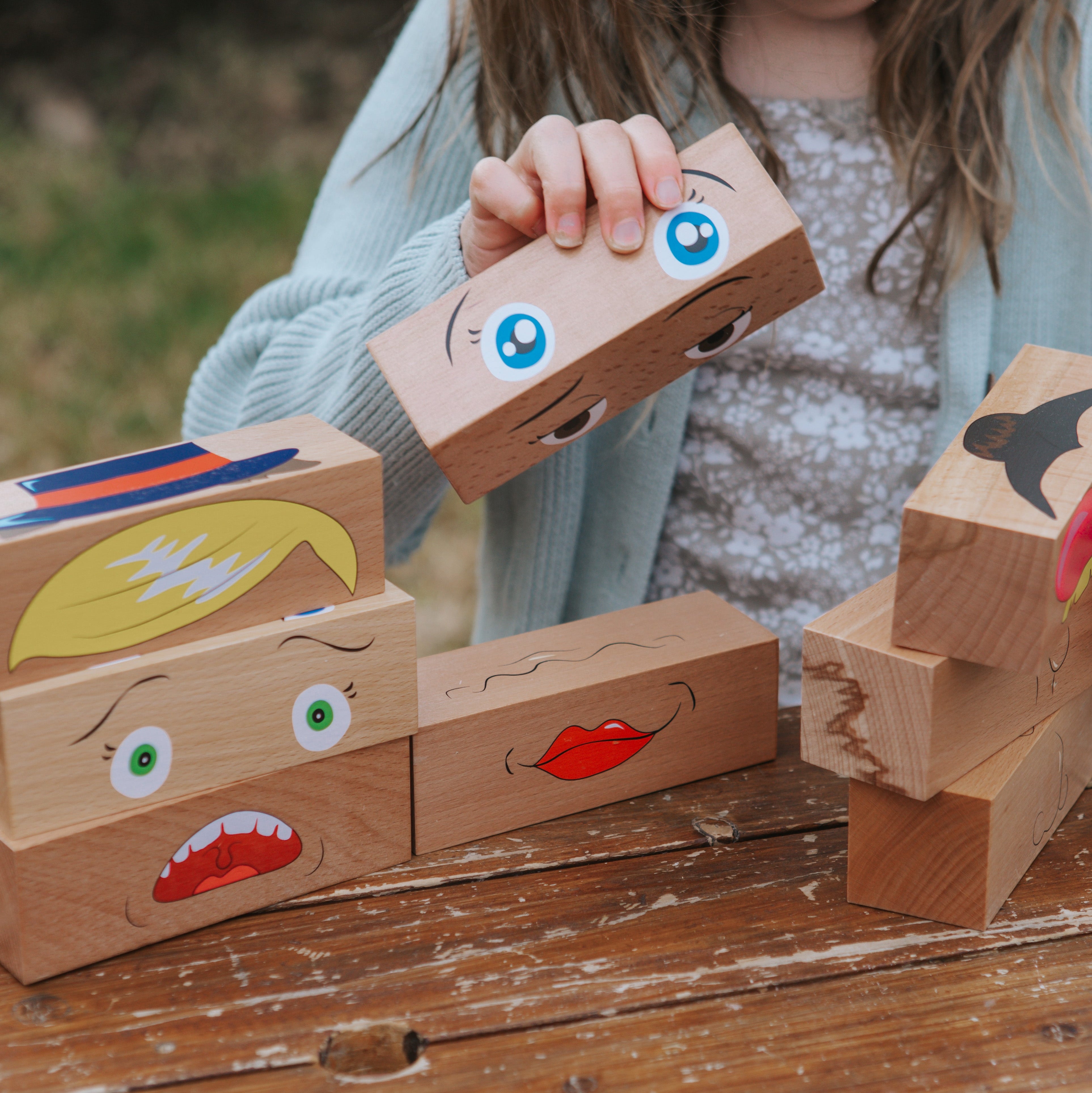 Child playing with wooden blocks with facial expressions on a wooden surface outdoors.